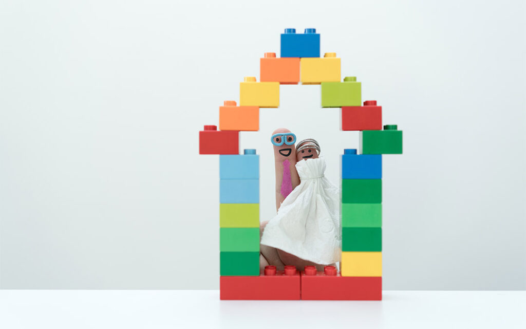 Photograph of two smiling finger puppets wearing wedding outfits standing inside of a building block church in a white space.