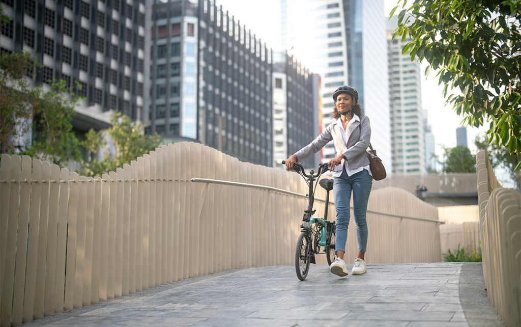 Photograph of a slightly smiling woman with a bicycle helmet on her head walking her bike down a side walk of a city.