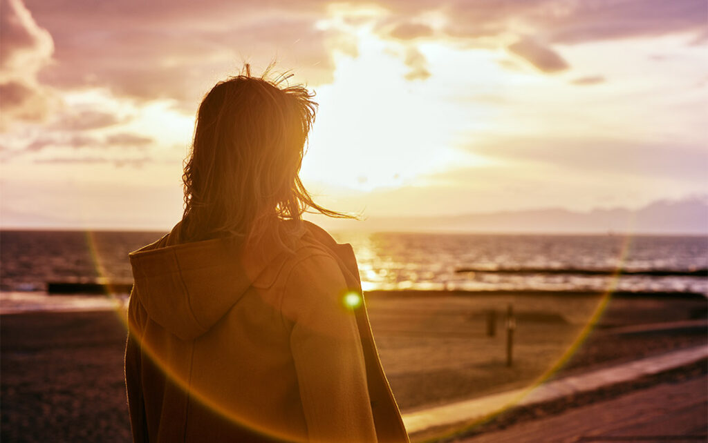 Photograph of a woman with a hooded jacket and slightly windblown hair looking into the distance at a beach with mountains in the distance and a sunsetting with clouds in the sky.