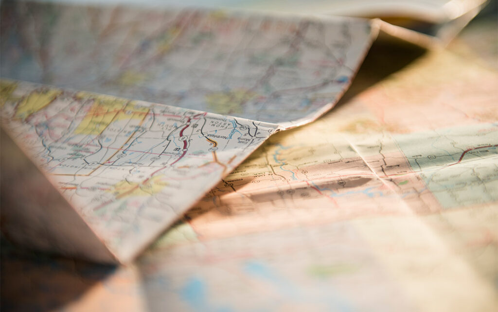 Closeup photograph of two road maps unfolded, with one laying on top of the other. The foreground and backgrounds are blurred white the middle ground is slightly more in focus but not sharp.