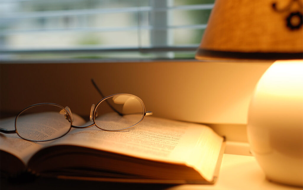 Photograph of eye glasses resting upside down on an open book on top of a table next to a table lamp that is turned on with a warm glow.