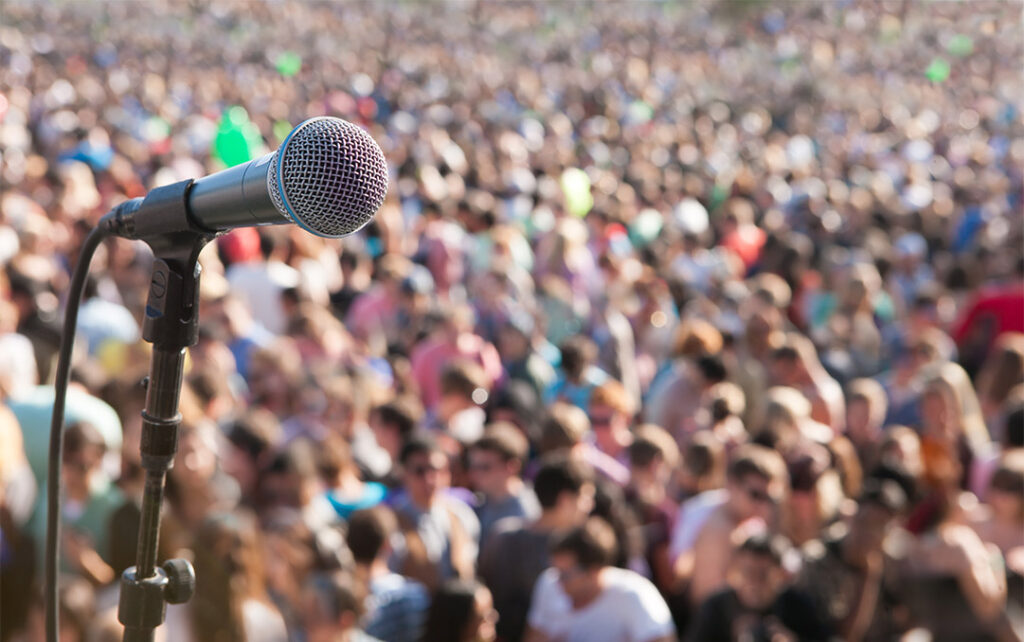 Photograph of a microphone on a stand in focus in the foreground of a sea of a blurred crowd of people.