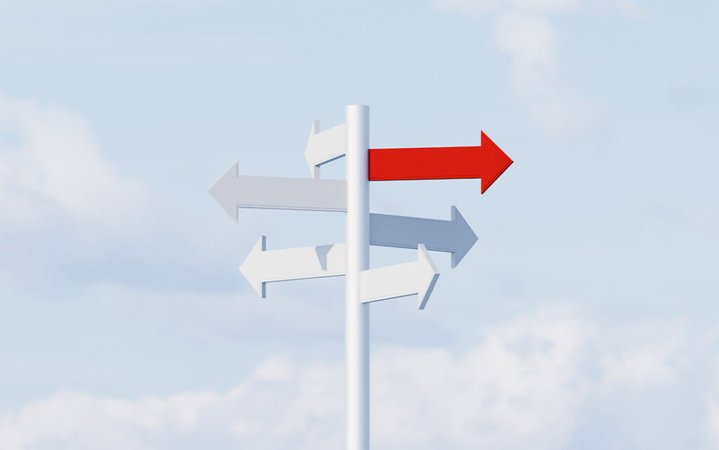 Photograph of a white pole with five white arrow shaped signs pointing in various directions. One of the arrow is red and points to the right and the background is a pale blue sky with clouds.