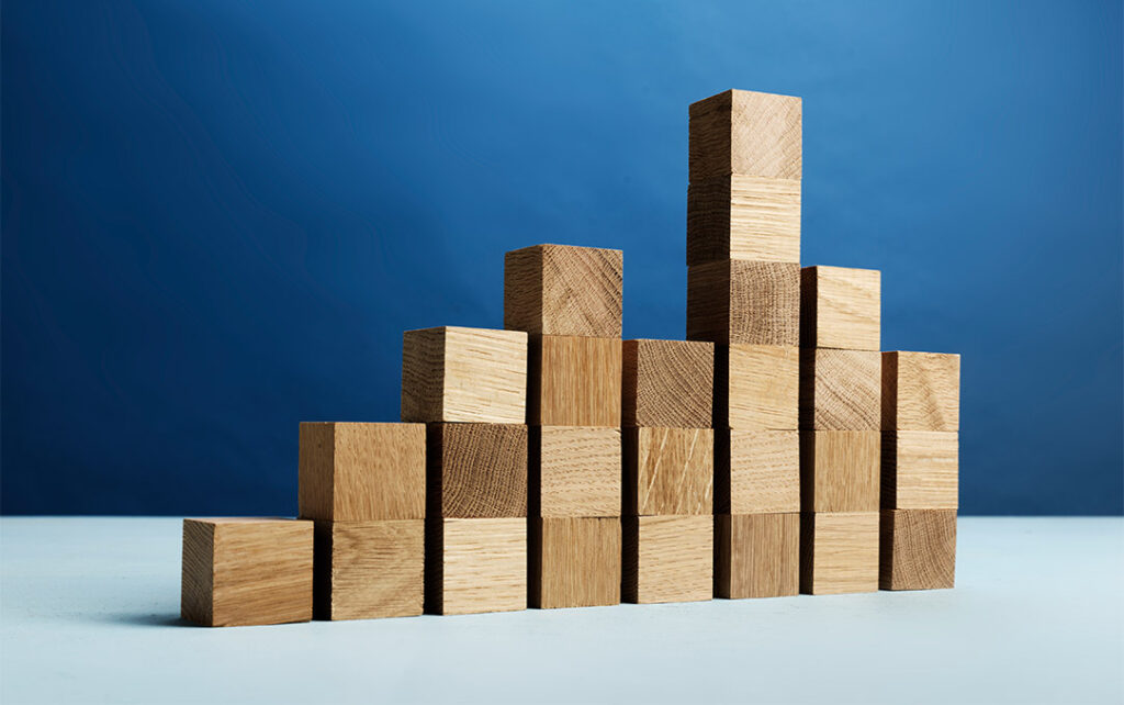 Photograph of eight stacks of square wooden blocks of various heights resting on a light blue surface with a darker blue wall behind it.