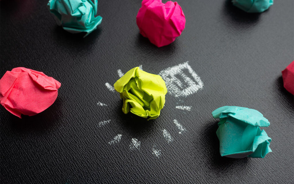 Closeup photograph of brightly colored bunched up post-it notes resting on a black surface. One of the post-its is bright yellow and has a broken white circle drawn in chalk on the black surface surrounding it.