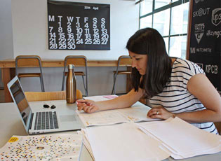 Woman in a striped shirt sitting at a table with an open laptop and a stack of papers. She is holding a pen. She is in an windowed office space with a chalboard of brand logos and a large calendar blurred in the background