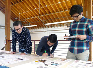 Three men standing over a table with pages of images and text spread out. They are all examining the images and two hold pens. One man is bent over writing on one of the images and another holds a stack of papers and is reading.