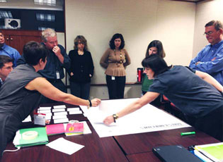 People in a conference room standing around a table with flashcards and a large poster laid on it. two women are slightly bent over the poster and point to highlight a specific point to the others in the room