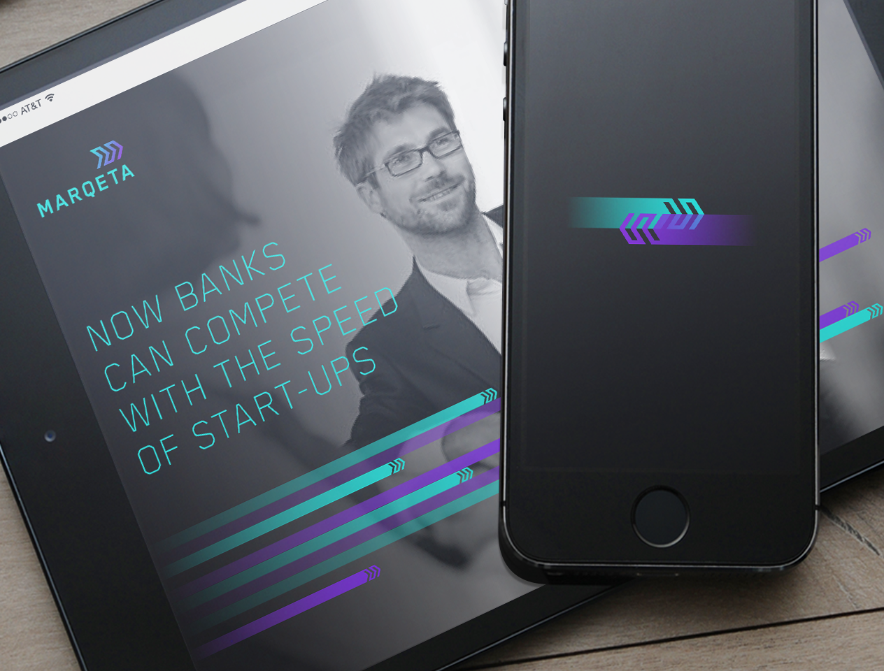 Photograph of a smart phone with the Marqeta outlined arrow logo in teal and purple suggesting motion, resting on top of a tablet. The tablet has the Marqeta website loaded which has a photograph of a smiling man with glasses in a suit. the text on the tablet says "Now banks can compete with the speed of start-ups"