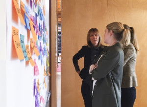 Three women in an office space looking at a wall of multi colored post-it notes. One woman has her arms crossed in front of her, another is just out of site and we see a partial profile. The third woman has her hand on her hips and is looking at the two other women with a serious expression on her face.