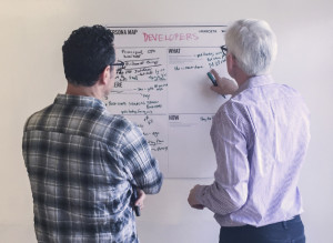 Photograph of two men looking at a white board with black writing on it. The man on the left has his arms crossed in front of him and the man on the right is pointing to something on the white board. The men have their backs facing us.