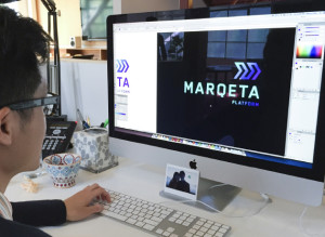 Photograph of a man with glasses sitting at a desk in an office working on a computer. The computer screen has a black rectangle with teal text that reads in all capital letters, "MARQETA" with smaller text underneath it that says in all capitals "PALTFORM" in a gradient teal, blue to purple font. On the top of the text above the "ETA" of the "MARQETA" is a gradient teal, blue, purple gradient double motion arrow pointing to the right.