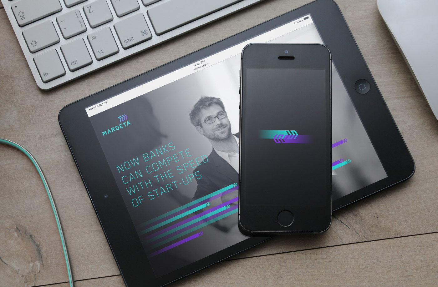 Photograph of a smart phone with the Marqeta outlined arrow logo in teal and purple suggesting motion, resting on top of a tablet. The tablet has the Marqeta website loaded which has a photograph of a smiling man with glasses in a suit. the text on the tablet says "Now banks can compete with the speed of start-ups"