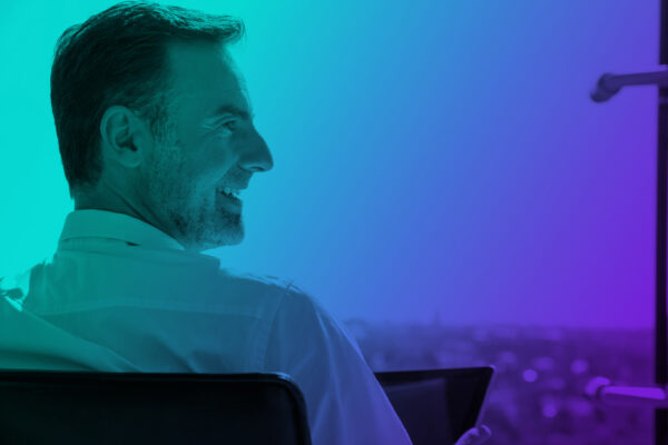 Rectangle of a photograph of the profile of a smiling man looking towards the right there is a teal, blue and purple gradient tone to the photo.