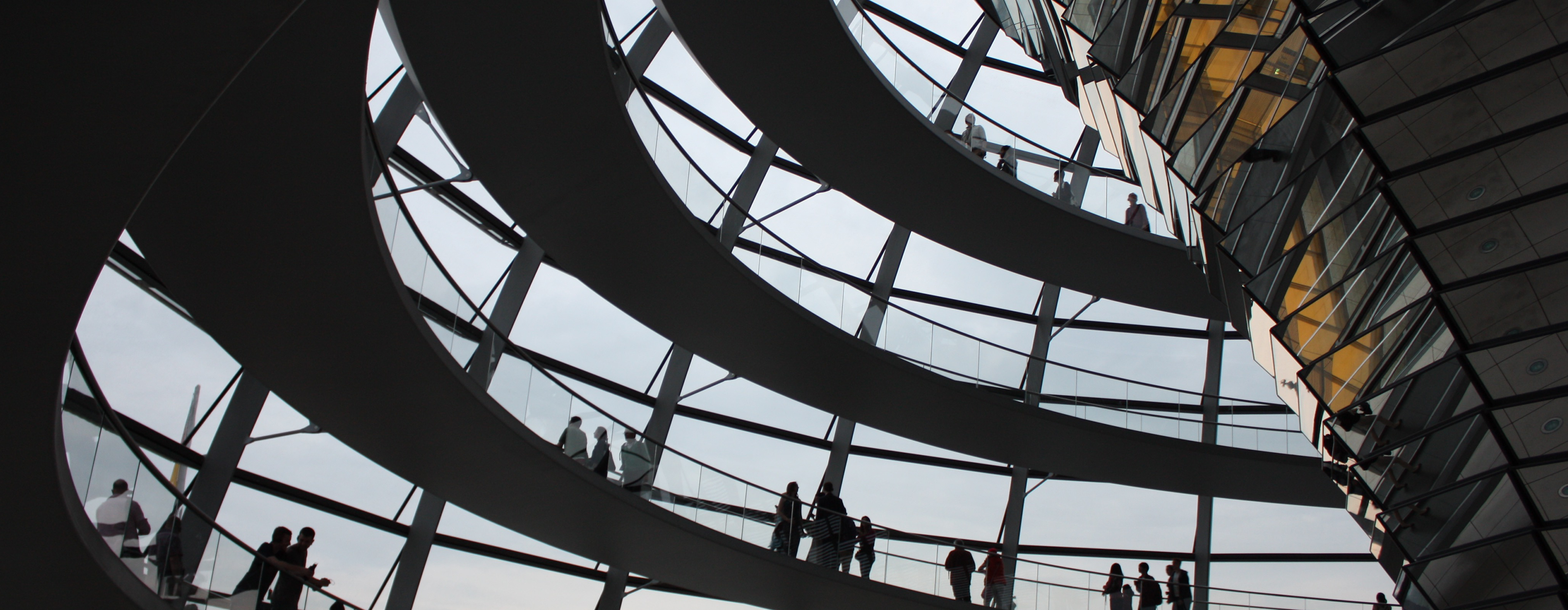 People standing on an indoor walkway of a building that spirals much like the Guggenheim Museum in New York City, looking out of the window at a monument with a blue sky