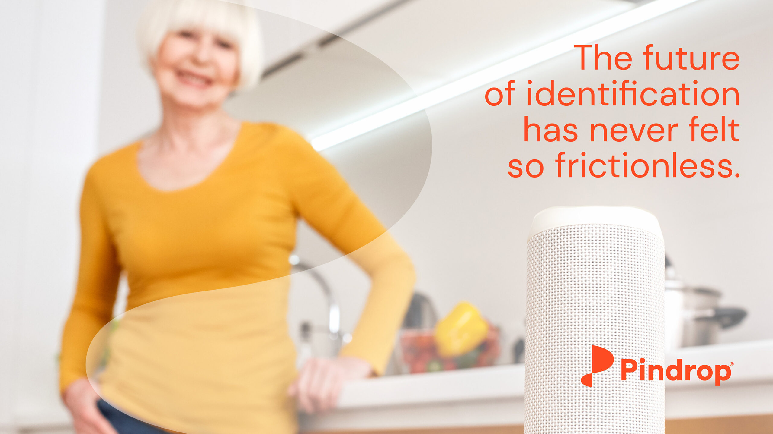 A blurred photograph of a an older woman in a yellow shirt standing in a white kitchen smiling at an Alexa type machine. Superimposed ontop of this is a light Pindrop logo with orange text that reads " The future of identification has never felt so frictionless." The Pindrop logo is on the bottom right corner of the image.