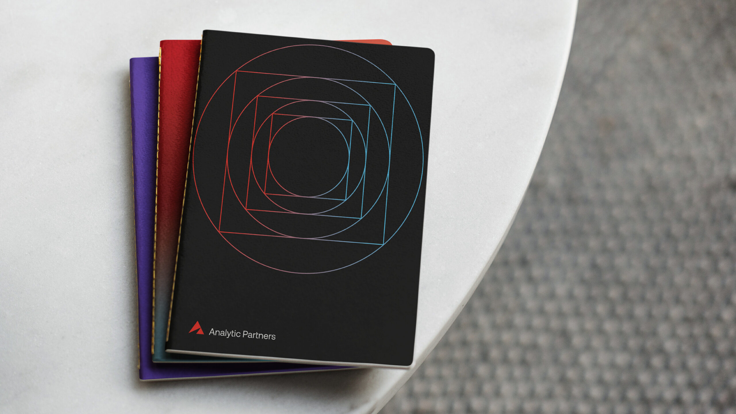 Photograph of three booklets stacked on top of each other sitting on a white table. The top book is black with a gradient red and blue outline of a circle inside of a square that repeats four times in the center of the booklet. White text that reads "Analytic Partners" with a red triangle on the bottom left corner. There is a purple booklet under the black booklet.