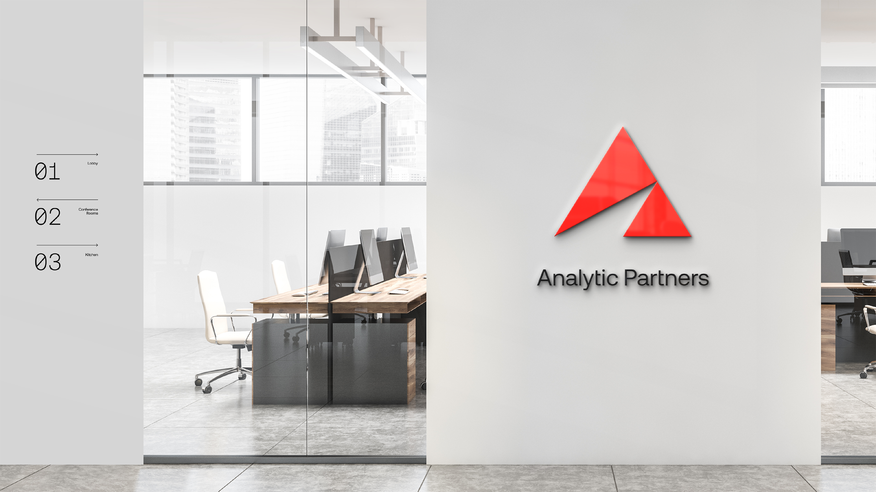 Photograph of a white office space with desks, computers and chairs. There is a white wall to the right of center with the Analytic Partners red triangle with a smaller diagonal triangle missing from its base logo and black text on the wall under the triangle that reads "Analytic Partners"