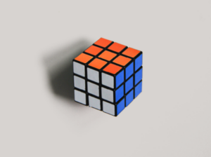 Photograph of a Rubics Cube in a white space. We see three sides of the cube which appears to be solved as all colors on each side match and are aligned. The top of the cube is Orange, the left side is white and the right side is blue.