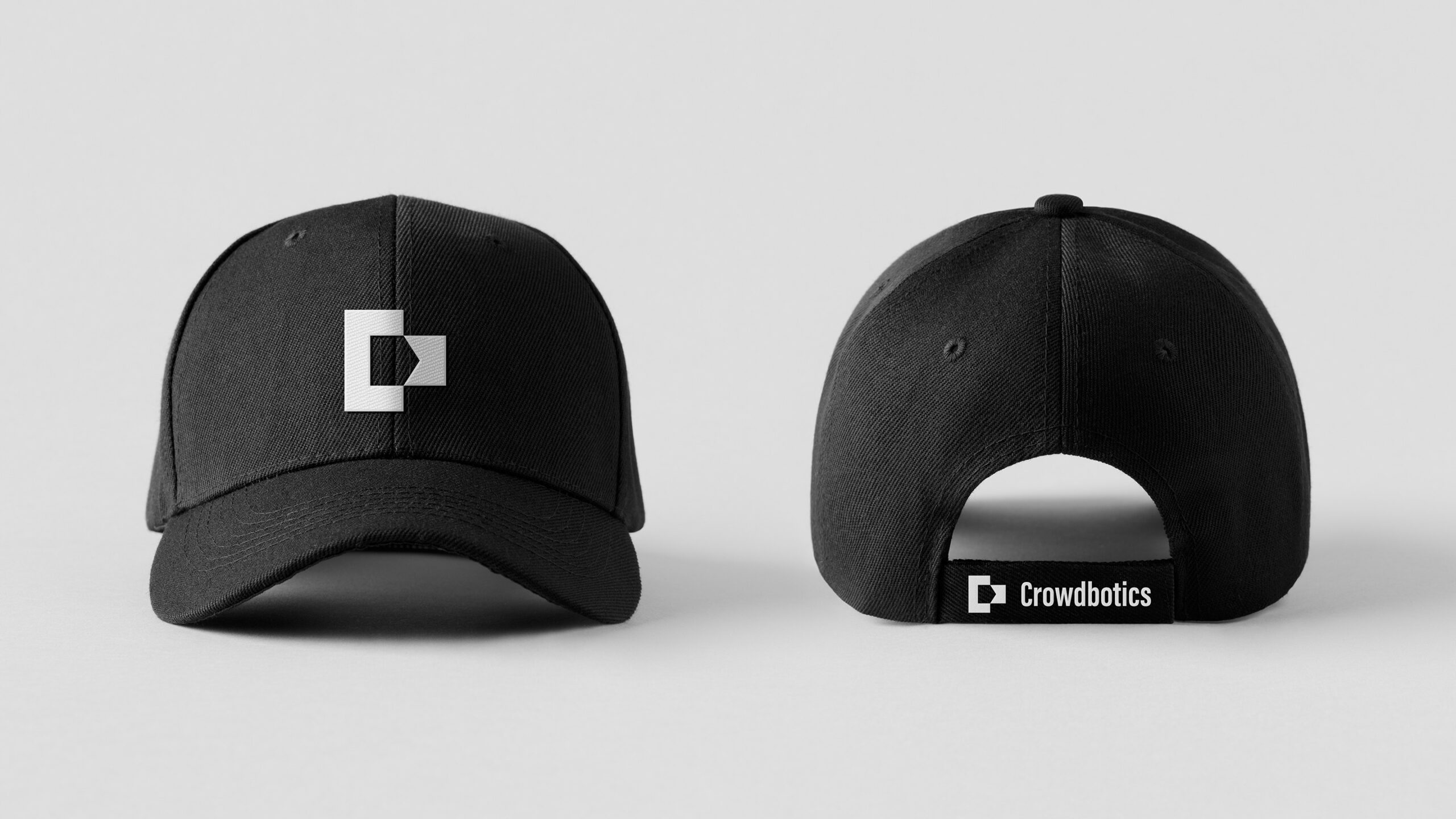 Photograph of two black baseball caps resting on a white surface in a white space. The left cap is facing the front and has the white Crowdbotics logo in the center, which is a half of a square "C" shape on the left with a smaller flag shape that touches the edge of the "C" with an inverted triangle that creates a negative space of a house on its side. The second hat in the right side is facing backwards and has a small white Crowdbotics logo with white text that says "Crowdbotics" on the hat's snaps.